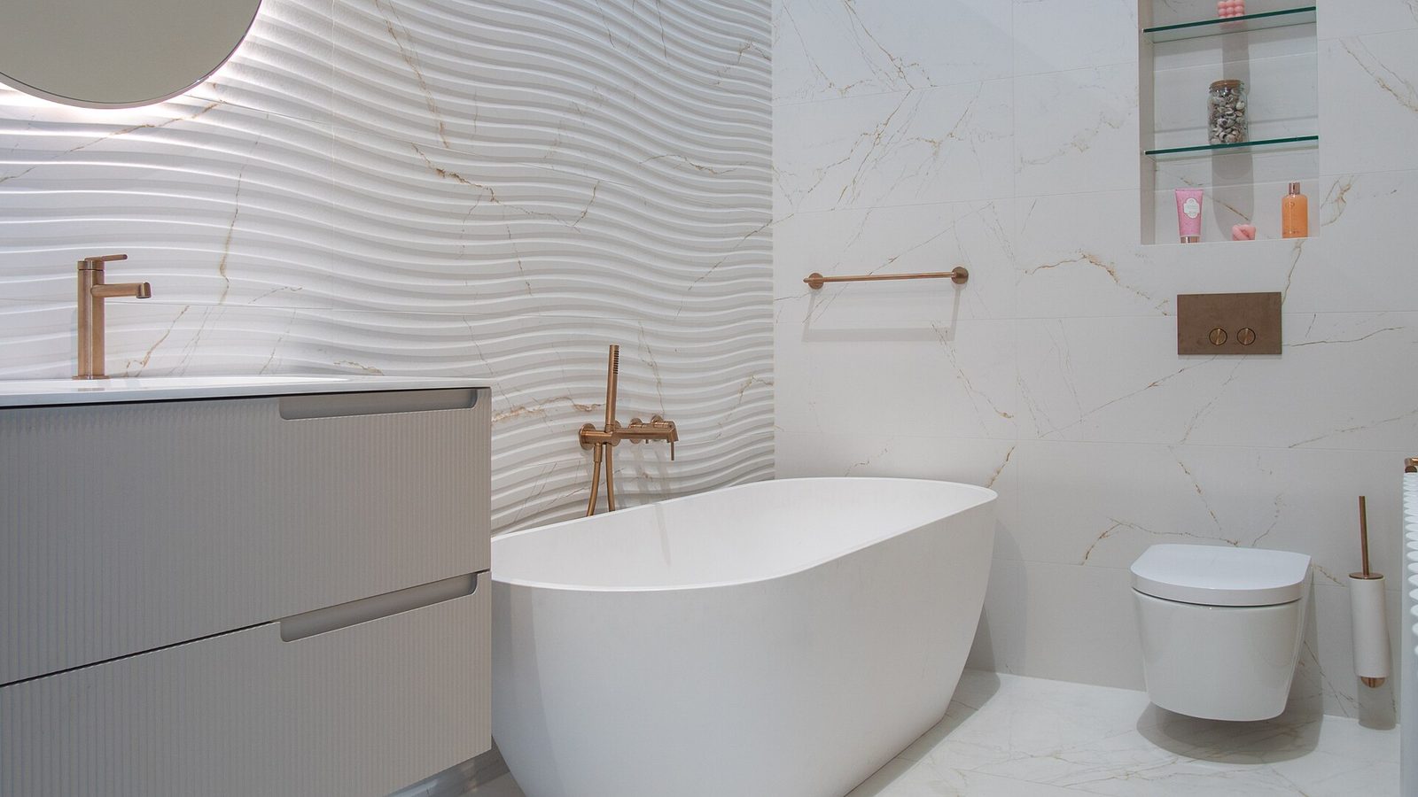 Badkamer met vrijstaand wit bad, marmerlook tegels, golvende wandtegels, grijs badmeubel en koperkleurige kranen