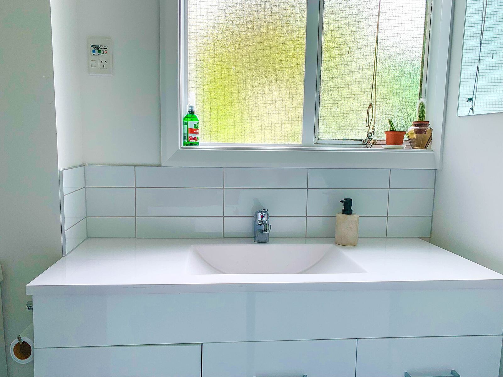 Wit wastafelmeubel met ingewerkte wasbak en witte wandtegels als spatwand onder een raam met matglas